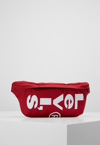 Marsupio rosso in tessuto con grandi lettere bianche. Presenta una chiusura con zip e una forma curva per un indossamento comodo. Superficie testurizzata.