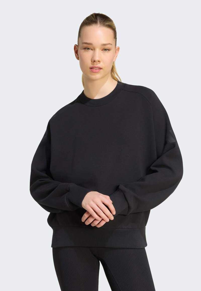 Jeune femme aux cheveux clairs attachés en queue de cheval, portant un sweat-shirt noir ample et un pantalon noir, debout devant un fond clair uni.