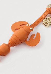 Oranžist nahast homaarikujuline võtmehoidja kuldse metallist klambri ja dekoratiivse riistvaraga valgel taustal.