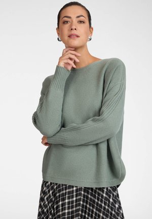 Mujer con cabello oscuro, suéter verde y falda a cuadros en blanco y negro, posando con una mano cerca del mentón y fondo neutro.