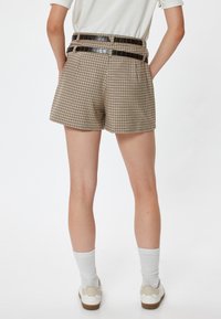 Shorts a cuadros en tan y marrón con una cintura alta, que incluyen un cinturón de cuero negro con relieve de cocodrilo y dos bolsillos traseros. Se muestran zapatillas blancas y calcetines.