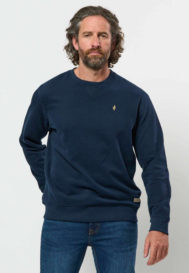 Homme aux cheveux bouclés et à la barbe portant un sweat bleu marine et un jean bleu sur un fond blanc uni.