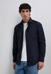 Camisa azul marino de botones con mangas largas, tejido suave y cuello estructurado, llevada sobre una camiseta blanca y combinada con vaqueros azules.