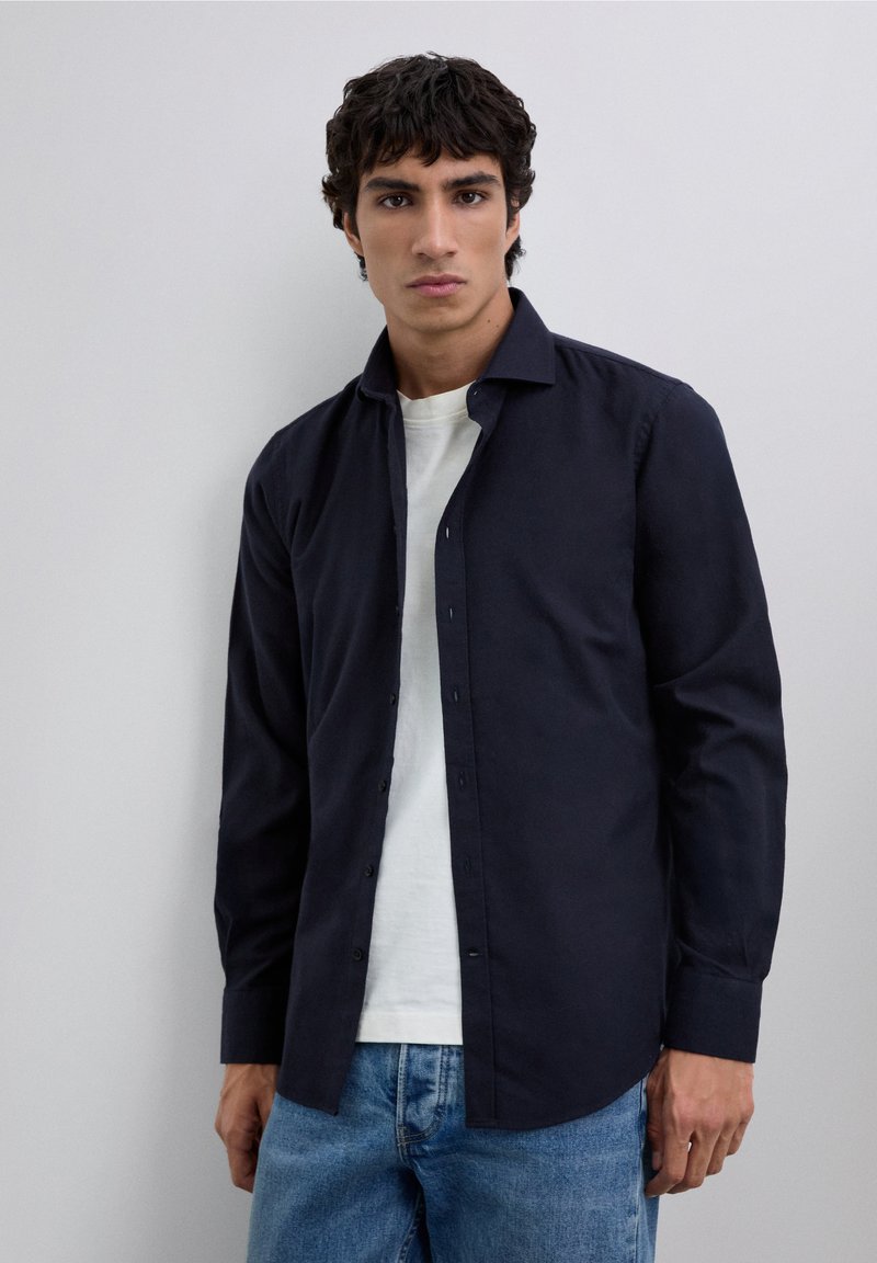 Camisa azul marino de botones con mangas largas, tejido suave y cuello estructurado, llevada sobre una camiseta blanca y combinada con vaqueros azules.
