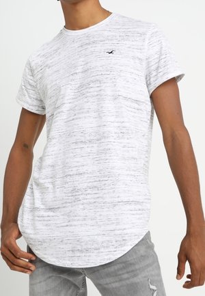 Man draagt een wit en grijs gespikkeld T-shirt met korte mouwen en een klein zwart logo op de borst, gecombineerd met lichtgrijze gescheurde spijkerbroek.