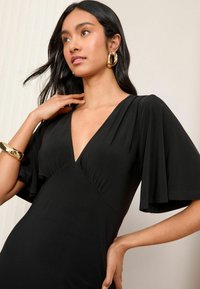 Rochie neagră cu decolteu adânc în V, mâneci fluturate și talie strânsă. Materialul pare neted și ușor. Accesorizată cu bijuterii din aur.