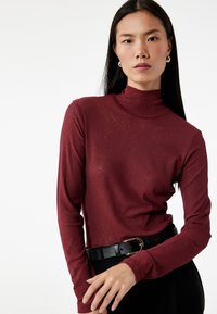 Bordeauxkleurige coltrui van een zachte, gestructureerde stof. Kenmerkt zich door lange mouwen en een slim fit ontwerp, gecombineerd met een zwarte riem met een gouden gesp.