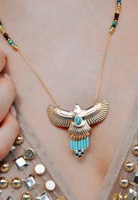 Collier en forme d'oiseau en or avec une pierre turquoise et des perles turquoise suspendues. La chaîne est ornée de perles colorées, le tout sur un fond neutre.