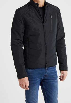 Homme portant une veste matelassée noire avec une poche poitrine zippée et un jean bleu sur fond uni.