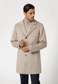 Beige vlněný kabát s přizpůsobeným střihem, se zářezovými klopami, zapínáním na jeden knoflík a bočními kapsami, nošený přes světře s vysokým límcem.