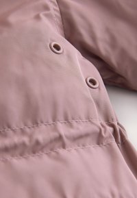 Gros plan d'un tissu mauve avec des œillets métalliques et des coutures cousues, montrant une partie de la texture et des détails de la surface d'un vêtement matelassé.
