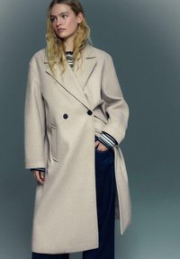 Svetlo bež volneni plašč z dvovrstno zasnovo, ki ima črne gumbe in stranski žep, nošen čez črtasto majico.