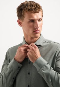 Groene overhemden met knopen, gemaakt van textiel met structuur, voorzien van een kraag, lange mouwen en donkere knopen, gedragen terwijl de handen de kraag aanpassen.