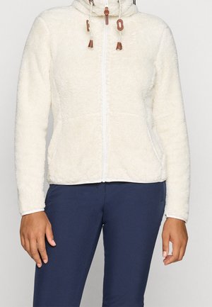 Personne portant une veste polaire blanche zippée avec des poches avant et un pantalon bleu marine, debout devant un fond uni.