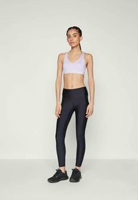 Világoslila sportmelltartó és fekete, magas derekú leggings rugalmas anyagból. Modell edzőcipőben egy semleges háttér előtt áll.