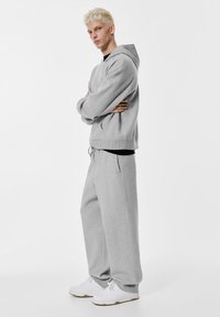 Šedá mikina s predným vreckom a kapucňou so šnúrkou, skombinovaná so širokými šedými teplákmi. Biely športový obuv dotvára vzhľad.