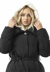 Schwarzer Steppmantel mit cremefarbenem Kapuzenfutter, großen Knöpfen, Taillenband und Steppmuster. Das Design ist locker geschnitten mit hohem Kragen.