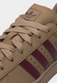 Smėlio spalvos Adidas sportbačiai su smėlio spalvos batraiščiais ir bordo dryželiais, ant liežuvėlio matomas Adidas logotipas ir šone užrašas „Superstar“.