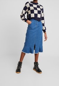 Jupe en denim avec un ourlet effiloché et une fente avant, associée à un pull à carreaux bleu marine et crème. Des bottes noires à lacets complètent le look.