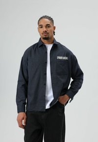 Zwart en marineblauw geruit overhemd met knopen en een borstzak waarop de tekst "FREE DOM" in het wit staat, gedragen over een wit T-shirt en zwarte broek.