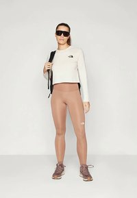 Svetle šedý zkrácený top s dlouhým rukávem a logem, béžové legíny s vysokým pasem, hnědé tenisky a sluneční brýle; štíhlý, přiléhavý design.