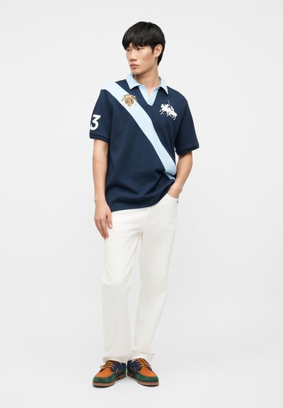 Άνδρας μοντέλο που φοράει μπλε-σκούρο πόλο με ανοιχτό μπλε διαγώνια ρίγα, λευκό παντελόνι και πολύχρωμα καφέ, πράσινα και μπλε κορδόνια παπούτσια, στέκεται με το ένα χέρι στην τσέπη.