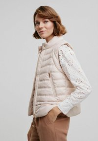 Jasnoróżowa kamizelka puchowa z pikowanym wzorem, z przodem na zamek błyskawiczny i wysokim kołnierzem. Nosi się ją na białej haftowanej bluzce z długim rękawem.