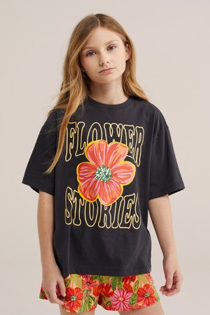 Mladé dievča s dlhými blond vlasmi nosí čierne oversize tričko s veľkou červenou kvetinou a nápisom "FLOWER STORIES", farebné kvetované šortky.