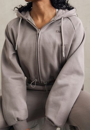 Personne portant un sweat à capuche mauve atténué avec fermeture éclair, cordons, taille élastique et pantalon assorti, assise jambes écartées.