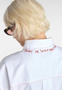 White shirt featuring red embroidery reading "TEMPT ME WITH A GOOD TIME" at the collar, with contrasting red stitching on the seams.