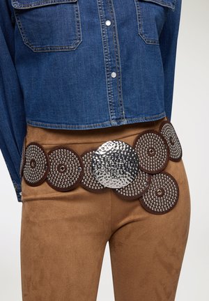 Cintura in camoscio marrone con grande centro in metallo argento martellato e dischi circolari in pelle marrone decorati con borchie argentate. Indossata sopra pantaloni color sabbia.
