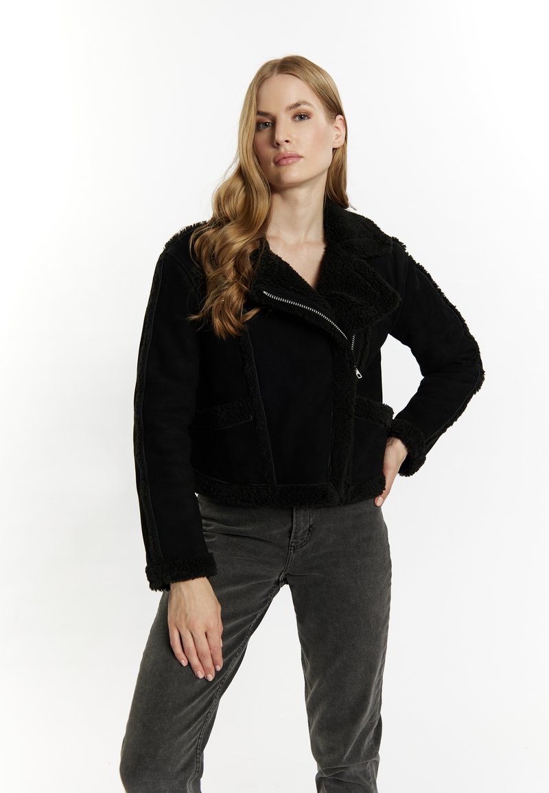 Schwarze Wildleder-Cropjacke mit großem Kragen und flauschigem Besatz. Verfügt über einen Reißverschluss und zwei Vordertaschen. Kombiniert mit grauen Jeans.