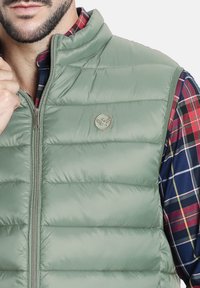 Chaleco acolchado verde con textura de saco horizontal, cuello alzado y insignia metálica de la marca en el lado izquierdo del pecho. Usado sobre una camisa de cuadros.