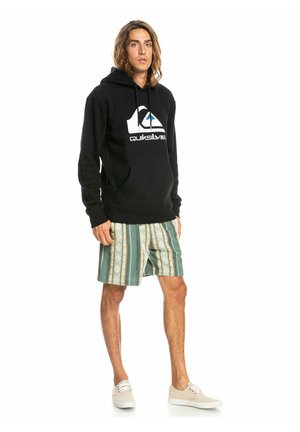 Sudadera negra con bolsillo frontal y logo, combinada con pantalones cortos a rayas en verde y beige. Modelo usando zapatos claros, de pie y erguido.