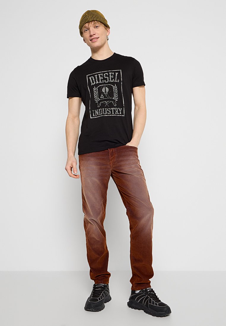 Un jeune homme portant un bonnet en tricot jaune, un t-shirt noir Diesel Industry, un jean marron et des baskets noires, se tenant devant un fond uni.