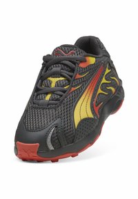 Sneaker nera in rete con accenti gialli e rossi a forma di fiamma. Presenta suola in gomma grigia e tomaia texturizzata con lacci per una calzata sicura.