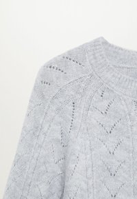Pull en tricot gris clair avec un col côtelé, présentant un motif texturé en chevrons et des mailles ouvertes, offrant une apparence douce.