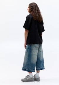 Fekete oversized póló, kék farmer culotte zokni, amely fokozatos mosással készült, és szürke sportcipő. Göndör haj és világos zoknik láthatók. Minimalista háttér.