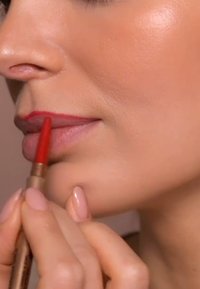 Red lip liner being applied to the lips, held by a hand. The lips are a natural shade, and the background is softly blurred.