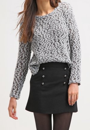 Femme portant un pull en tricot gris texturé rentré dans une mini-jupe noire ornée de boutons argentés décoratifs et des collants noirs transparents.