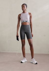 Sportbh i lyserød, højtaljede mørke shorts og hvide sneakers. Holder et sjippetov, stående på en neutral farvet gulv.