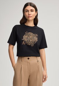 T-shirt noir à manches courtes et coupe ample, avec un design de logo circulaire doré et des motifs sur le devant. Texture en coton doux.