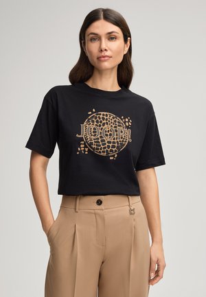 Zwarte T-shirt met korte mouwen en een losse pasvorm, voorzien van een cirkelvormig goudkleurig logoontwerp met patronen op de voorkant. Zachte katoenen textuur.