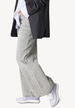 Person wearing light gray wide-leg jeans, white-purple sneakers, a white shirt, and a black blazer with one hand in pocket.