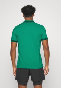 Zielona koszulka polo z klasycznym kołnierzykiem, z czarnymi paskami na rękawach. Miękka, gładka tkanina; dopasowany krój, pokazany widok z tyłu.