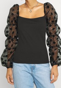 Top nero con scollatura quadrata e maniche a palloncino trasparenti con pois neri. Indossato con jeans azzurri chiari. Collana in argento visibile.