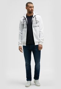 Sudadera blanca con cremallera y gráficos de texto negro, llevada sobre una camiseta oscura, combinada con jeans oscuros y zapatillas blancas. Textura de tela suave.