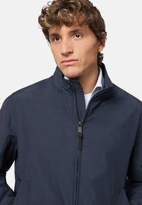 Giacca leggera blu navale con colletto alto, chiusura frontale con zip e tasche laterali; trama liscia e design minimalista.