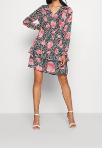Abito floreale con maniche lunghe, scollatura a V, orlo a strati con volant, fondo nero con rose rosa e foglie verdi, realizzato in tessuto leggero.