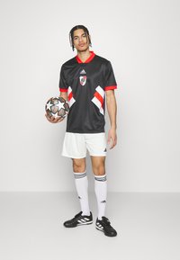 Jersey negro de Adidas con acentos y rayas rojas, pantalones cortos blancos, botas negras, sosteniendo un balón de fútbol con un diseño, con el logotipo del equipo de manera prominente.